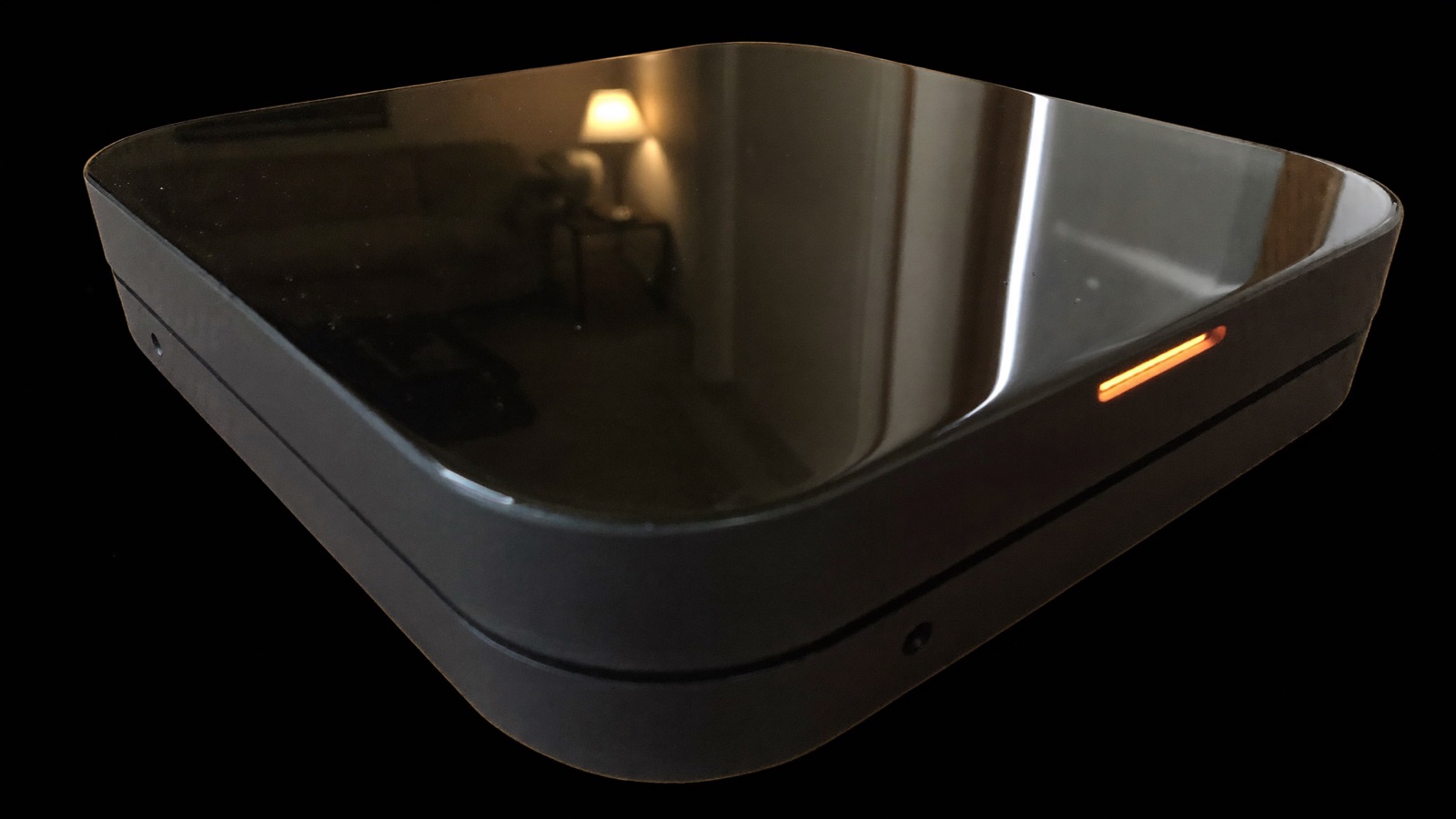 Close up across the glossy black glass top of the Glassback box, a living room reflected in it and one small amber light glowing at the edge.